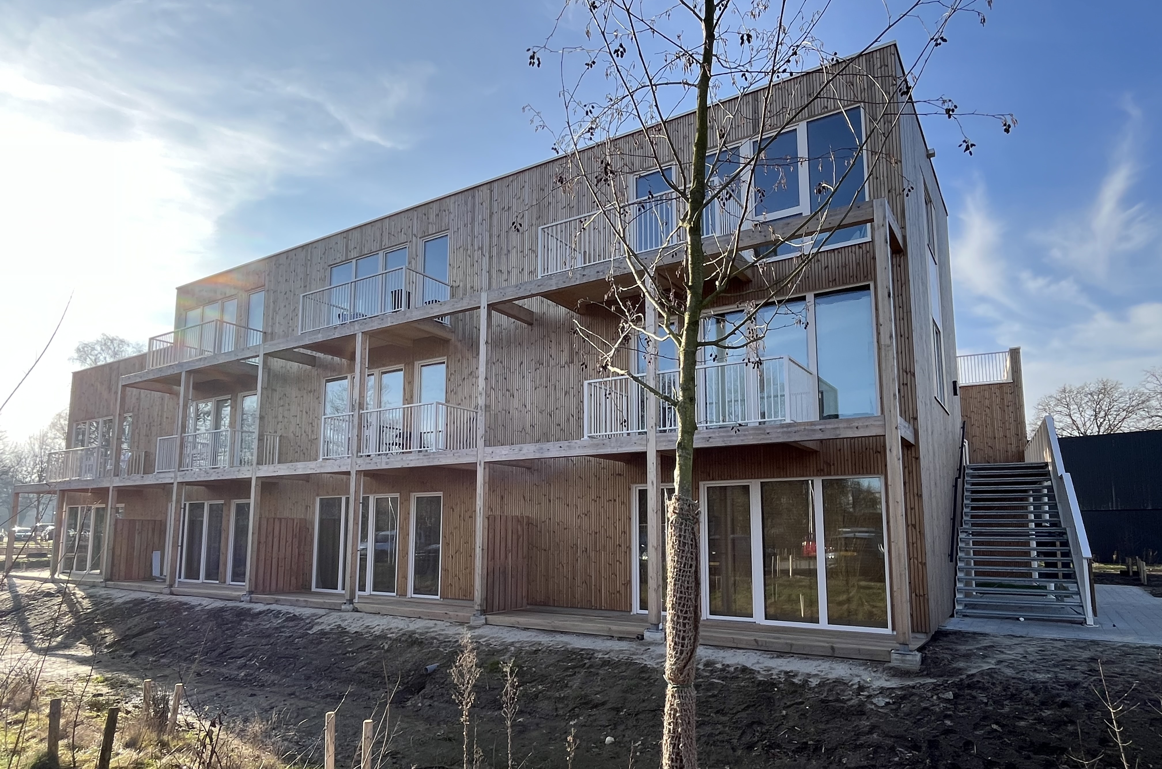 Modern houten gebouw met twee verdiepingen en glazen deuren, een buitentrap rechts en een kale boom ervoor onder een blauwe lucht.
