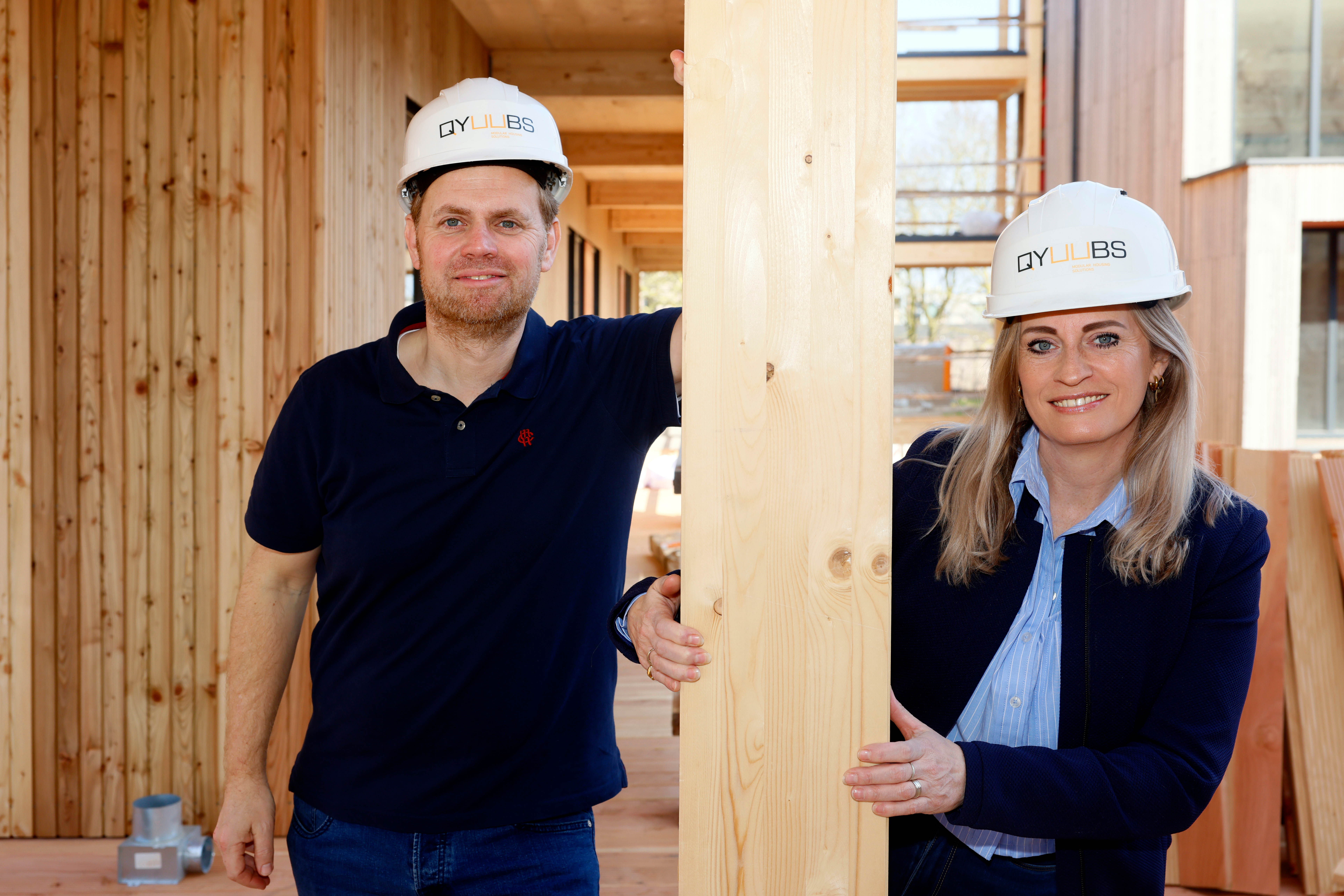 Twee bouwprofessionals met veiligheidshelmen staan naast een houten balk op een bouwplaats.