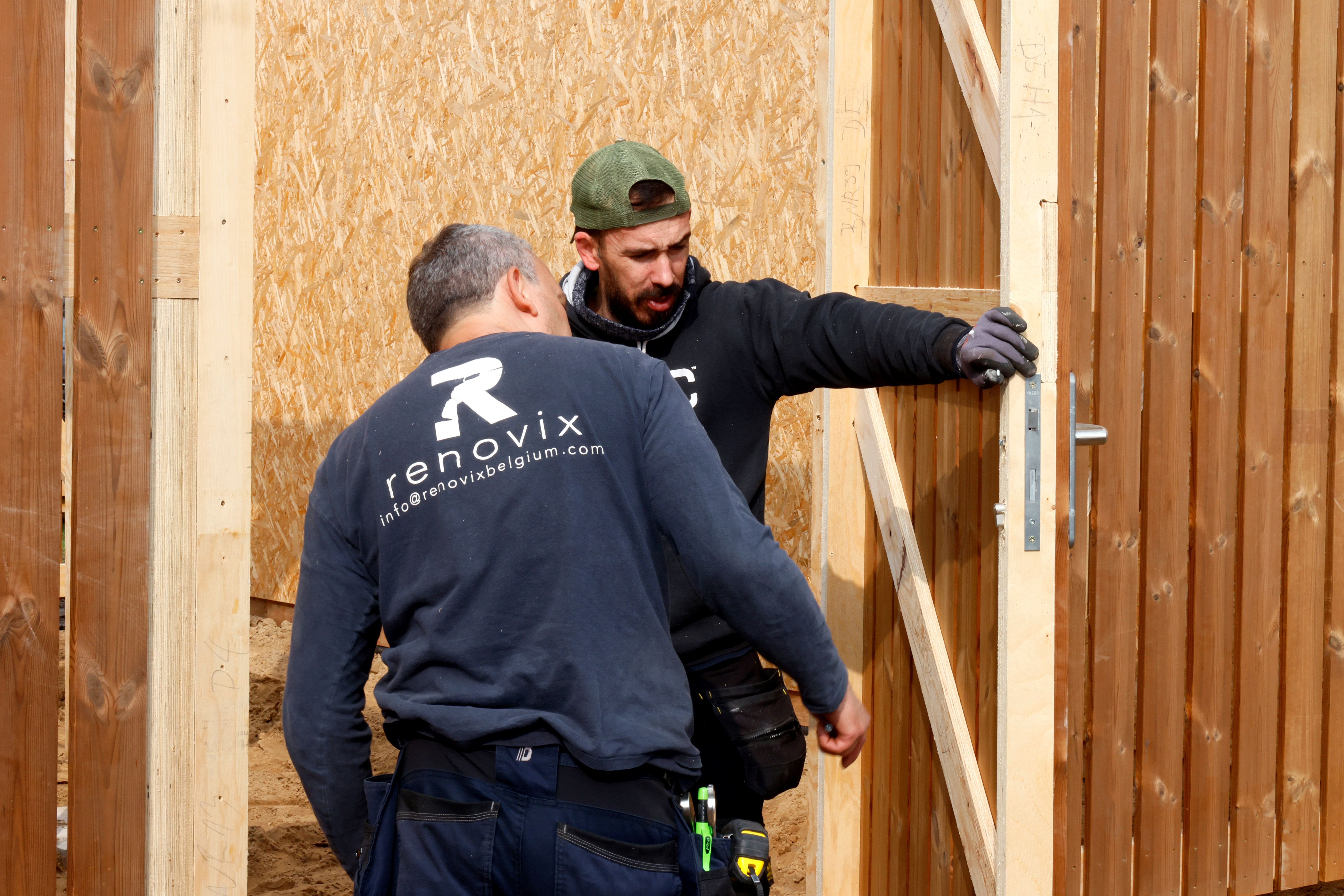 Twee bouwvakkers plaatsen een houten deur in een kozijn buiten.