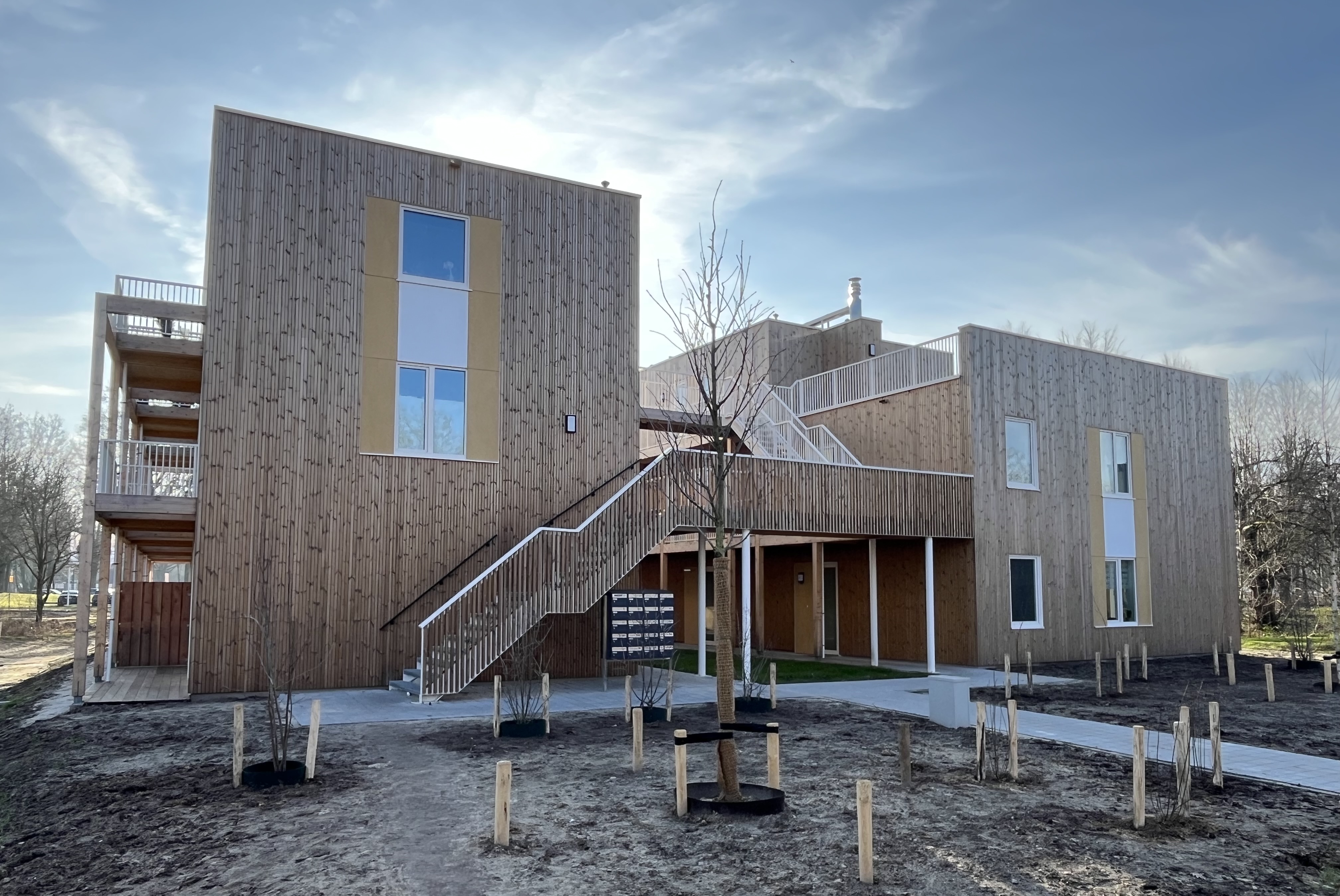 Modern houten appartementencomplex met buitentrap en pas geplantte bomen onder een blauwe lucht.