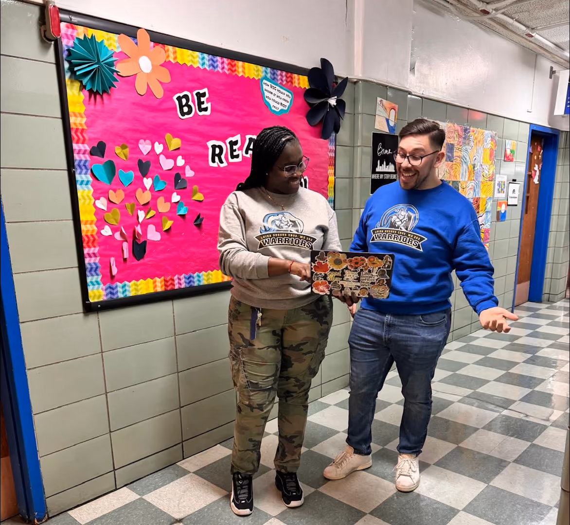 Two Warriors staff members smiling while sharing a decorated plaque in school hallway