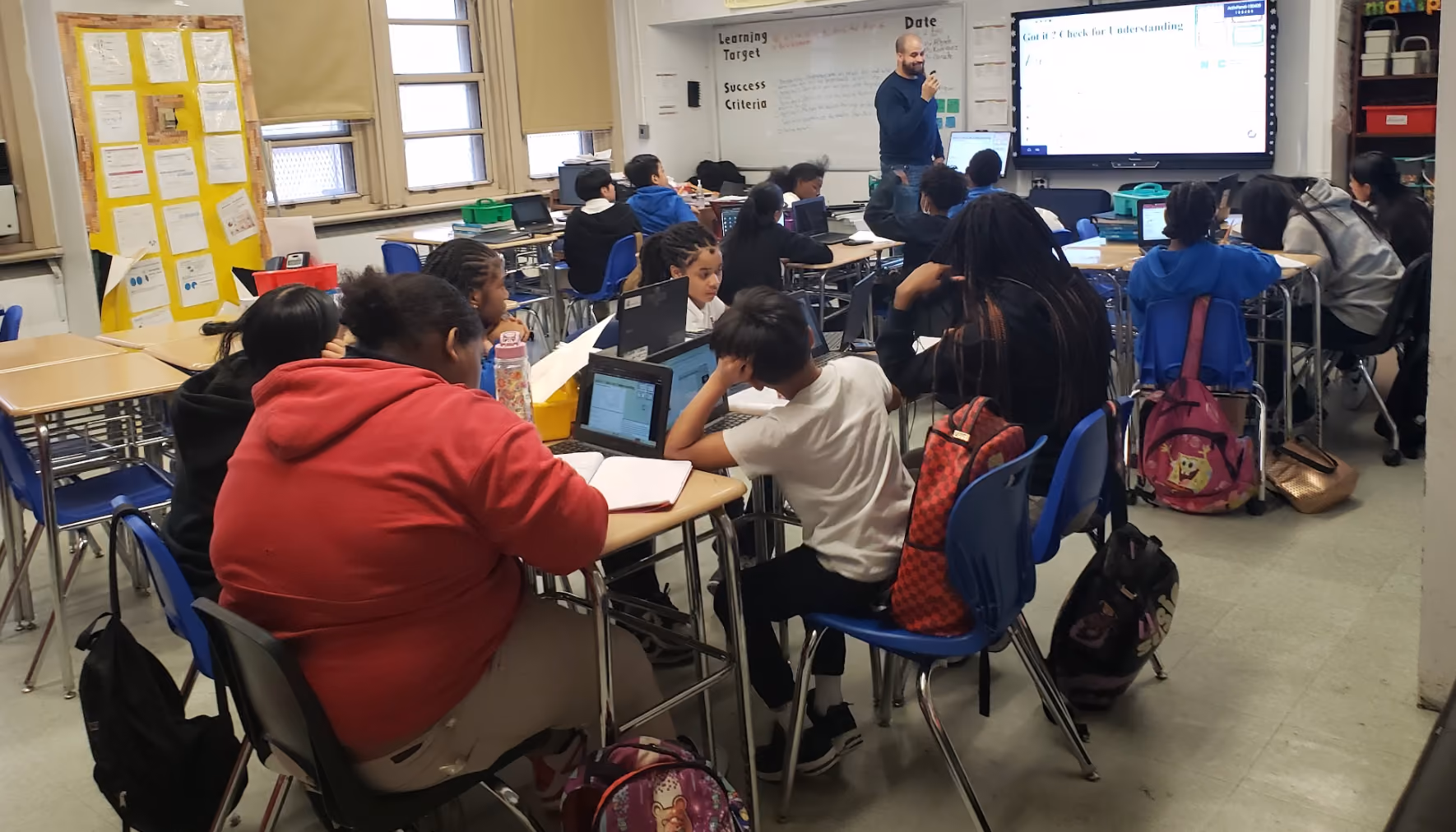 Teacher leads a lesson while students work on laptops in classroom