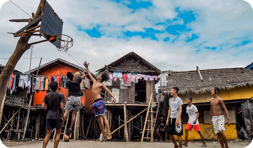 A group of young men in a third-world country playing basketball in front of some make-shift houses.