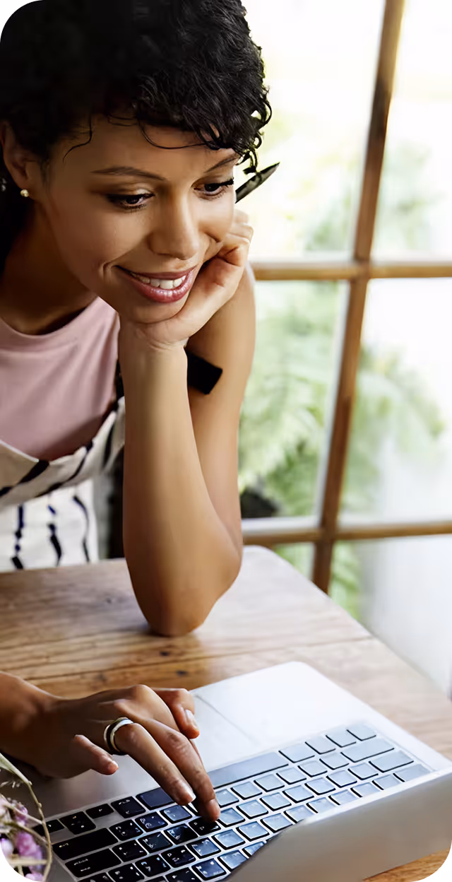 Mulher sorridente usando laptop em uma mesa de madeira perto de uma janela com plantas.