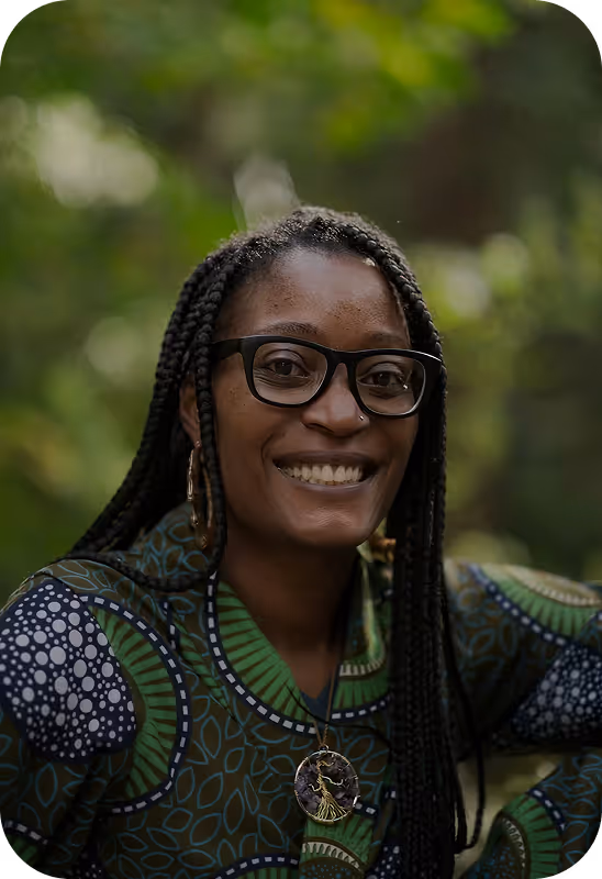 Mulher sorridente com tranças e óculos, usando blusa estampada e colar com pingente, em fundo de natureza desfocado.