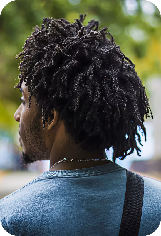 Homem visto de costas com cabelos estilo dreadlocks e camiseta cinza em ambiente ao ar livre com fundo desfocado.