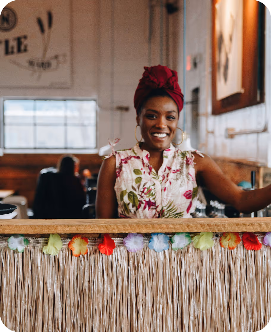 Mulher sorridente usando turbante vermelho e blusa florida atrás de um balcão decorado com flores tropicais.