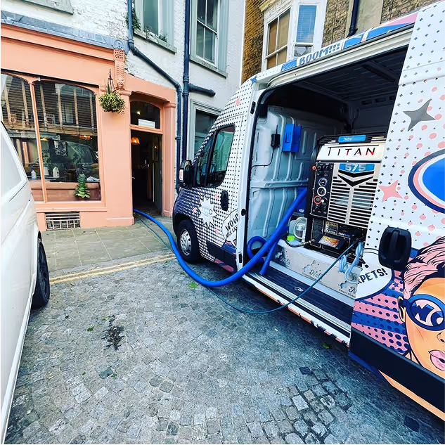 Painted van with cleaning equipment and blue hoses parked on a cobblestone street outside a peach-coloured building.