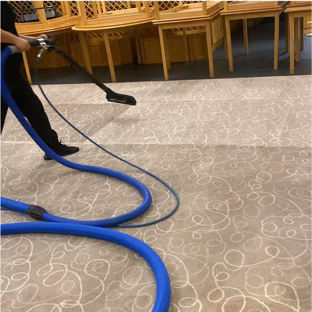 Person cleaning a beige carpet with white swirl patterns using a large blue hose and carpet cleaning tool in a room with wooden chairs stacked against the wall.