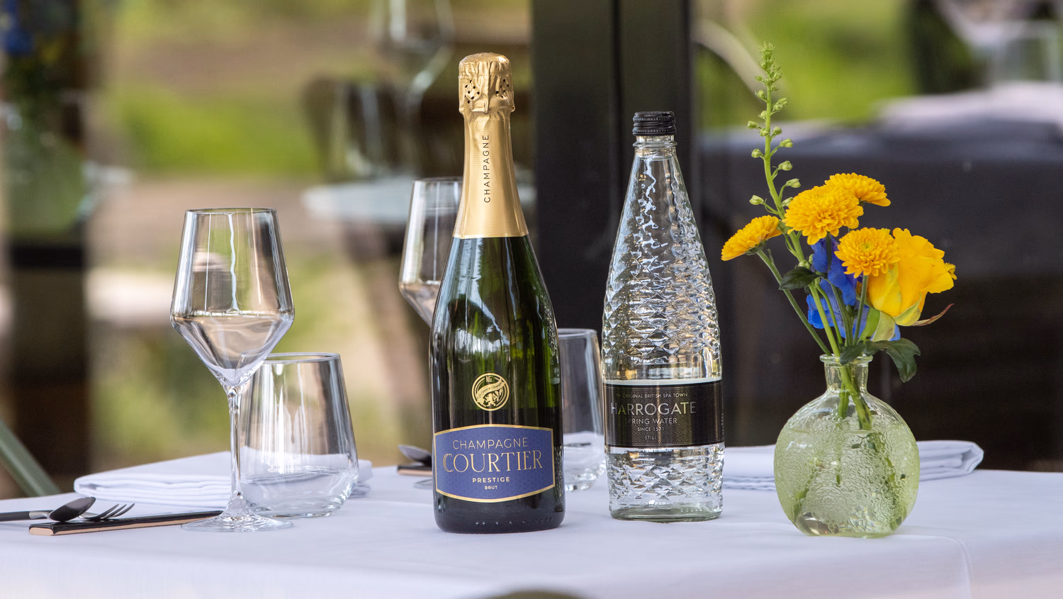 Tafel met fles champagne, fles mineraalwater, glazen en een vaas met gele bloemen.