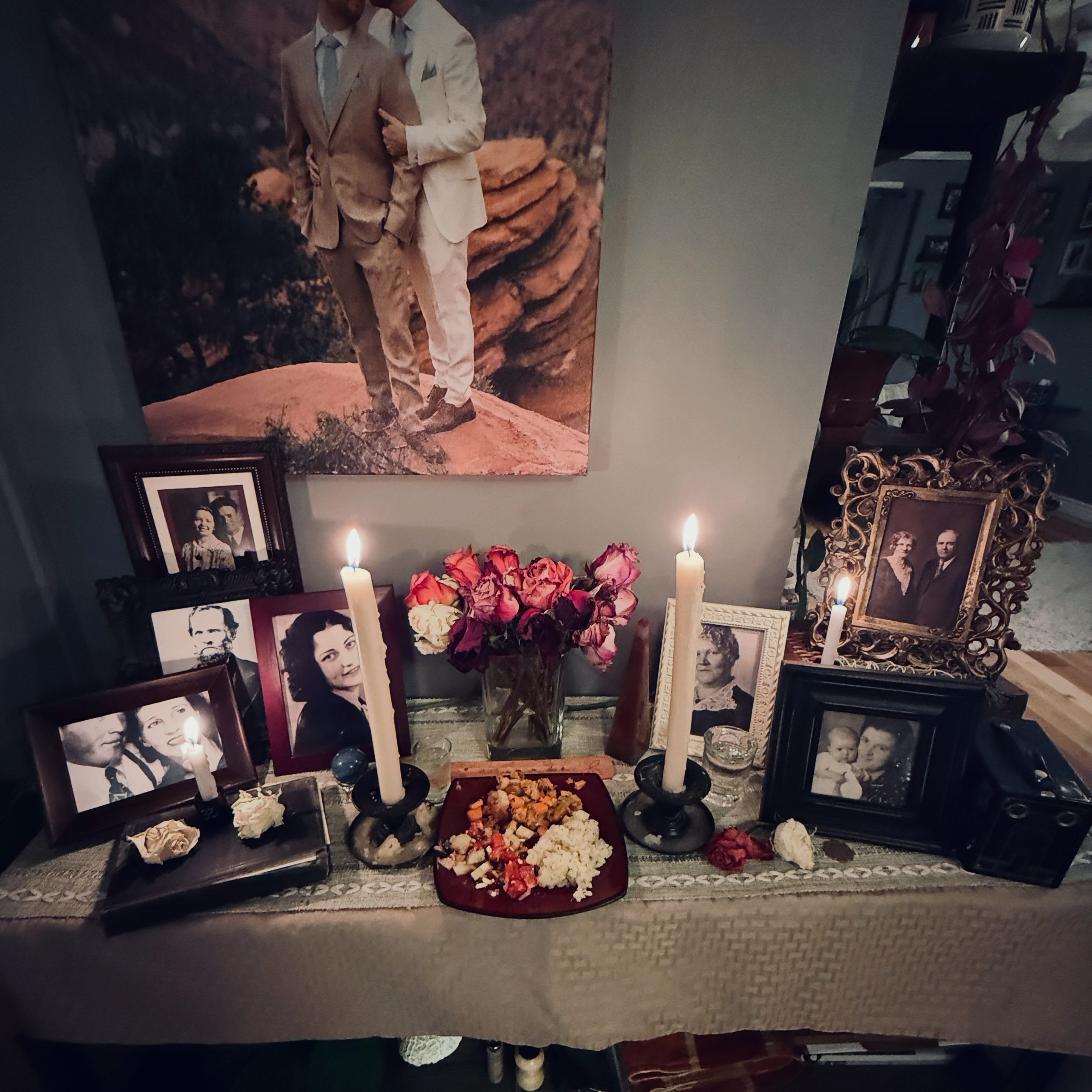 Candlelit ancestor altar for Samhain with framed family photos, lit candles, dried roses, and a plate of food offerings on a lace-covered table beneath a canvas photo of two men in suits.