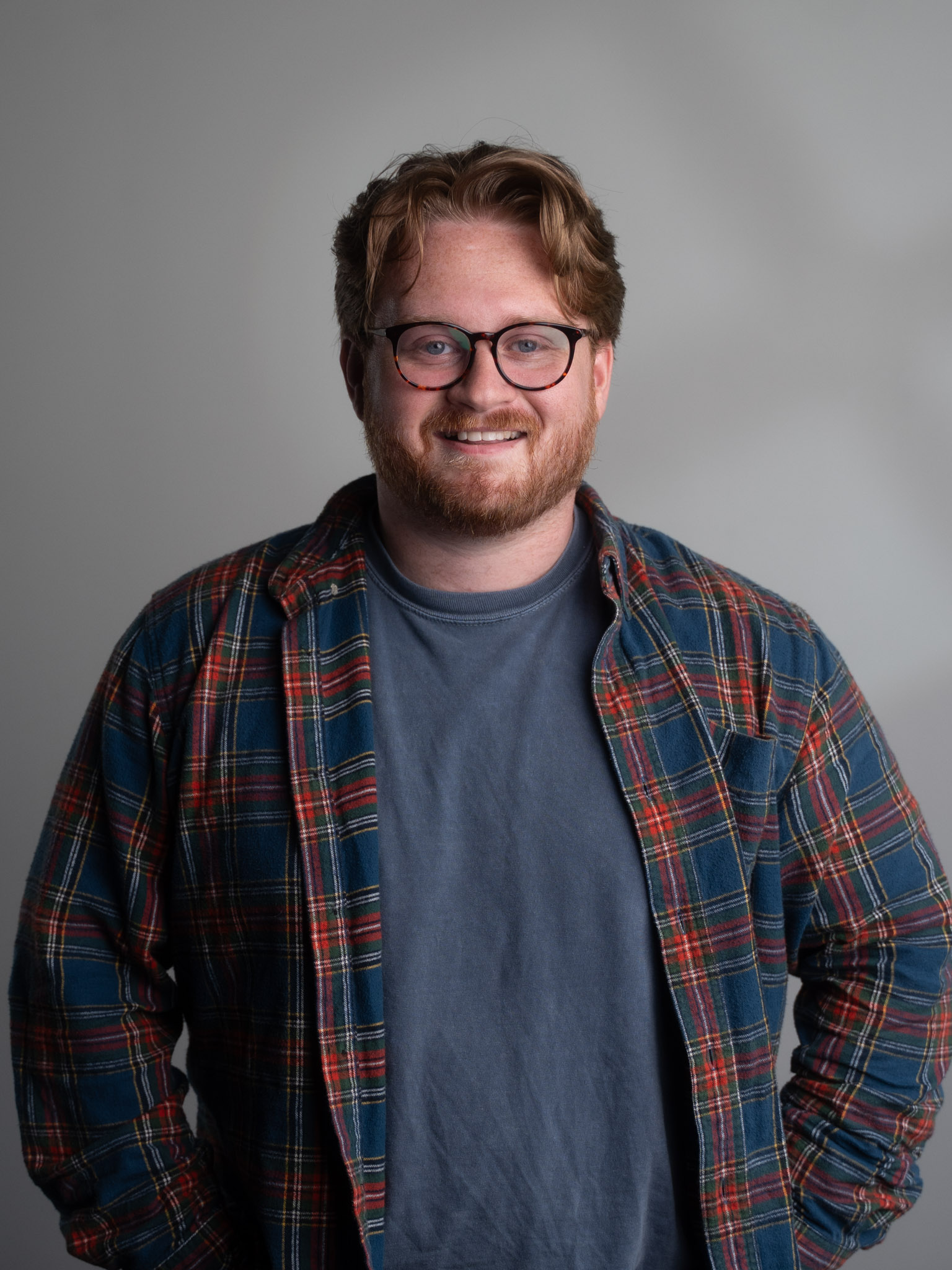 Smiling man with glasses and red hair wearing a blue and red plaid shirt over a gray t-shirt.