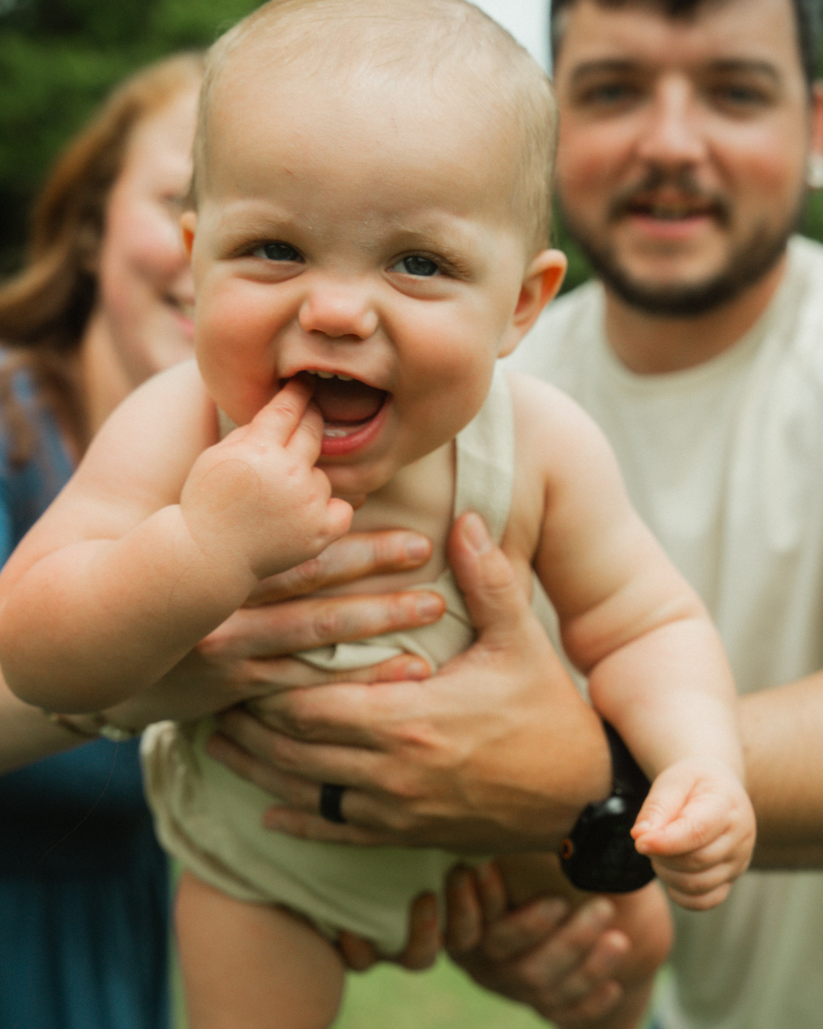 Smiling baby with fingers in mouth held by adults in outdoor setting.