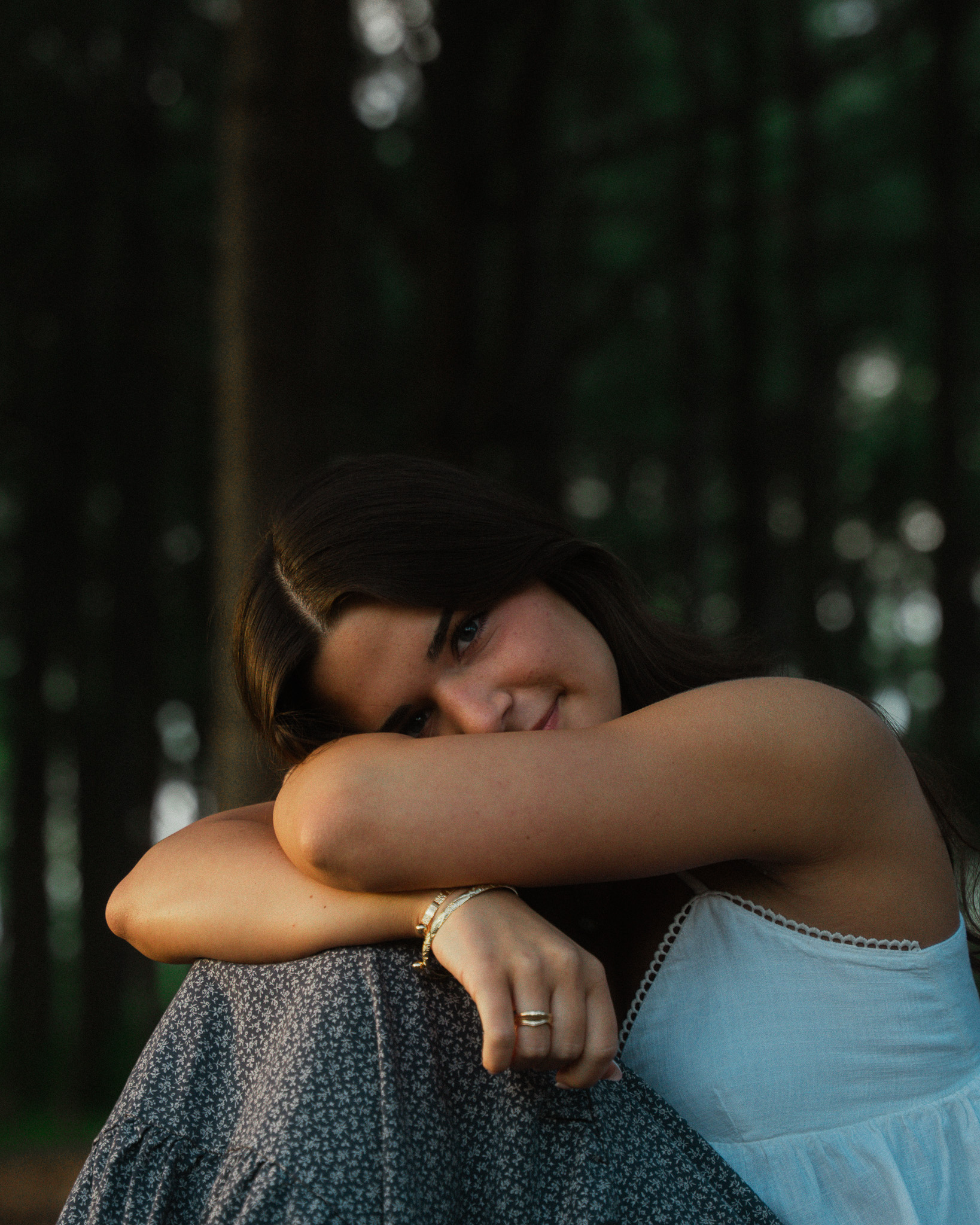 Young woman with dark hair resting her arms on her knees, smiling softly in a wooded outdoor setting.