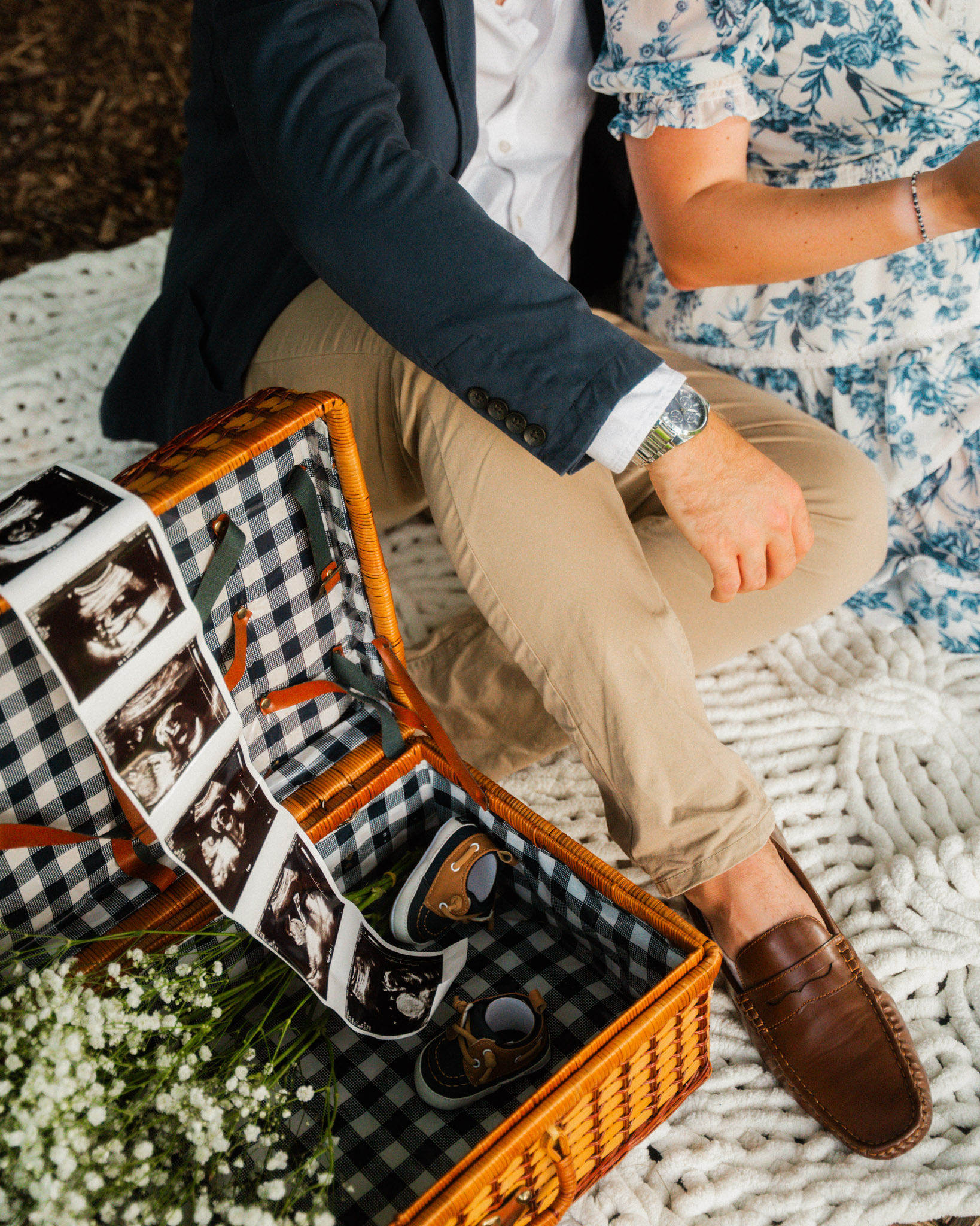 Couple sitting on a blanket next to an open picnic basket holding baby shoes and ultrasound images.