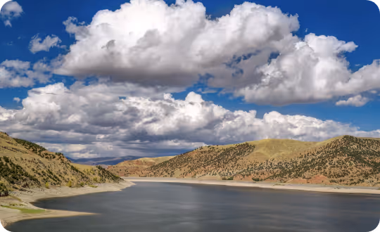 A serene, placid lake surrounded by a landscape of arid mountains, all under a vast blue sky filled with large, fluffy clouds.