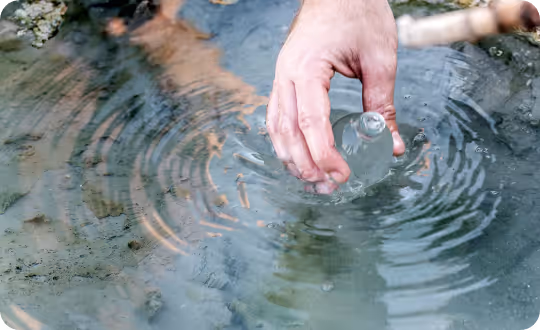 A hand holds a small, clear water bottle, filled with liquid, partially submerged in a body of water, likely collecting a research sample.