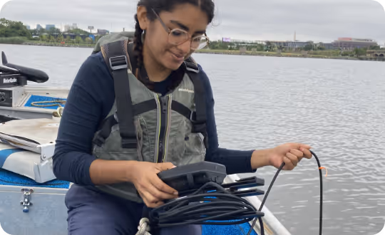 A researcher in a small boat on a tranquil lake holds scientific equipment for testing water quality.