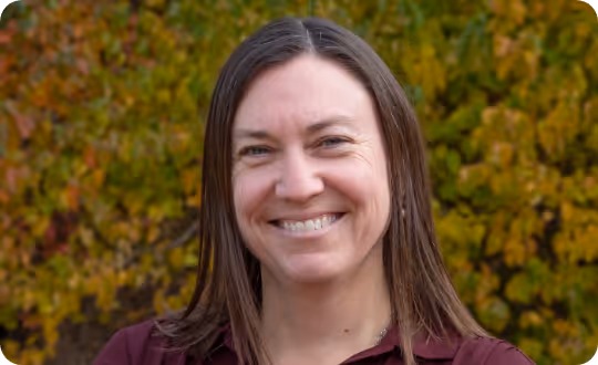 A close-up photograph of Professor Kendra Spence Cheruvelil smiling, framed by a soft-focus backdrop of vibrant foliage.