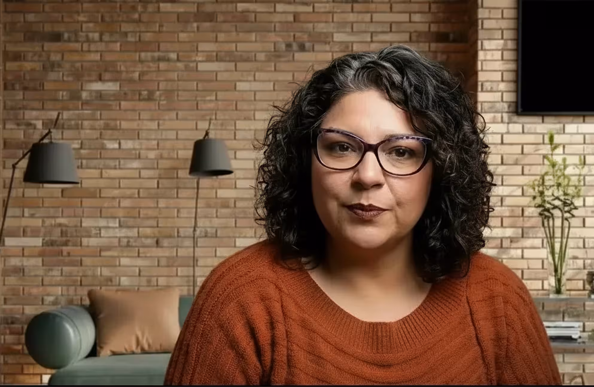 Woman with curly dark hair and glasses wearing an orange sweater sitting in a room with a brick wall, floor lamps, a sofa with a beige pillow, and plants.