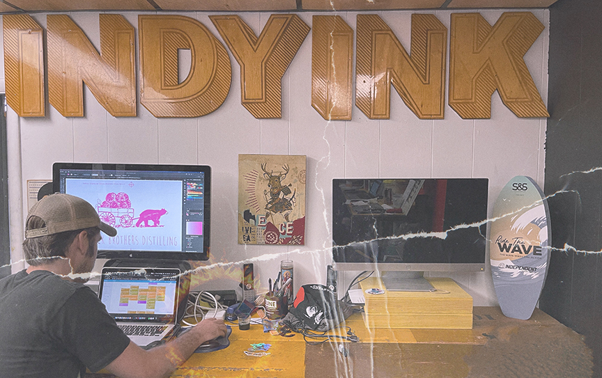 Designer working at a cluttered desk under large wooden “INDYINK” signage, creating artwork on a monitor using Adobe Illustrator. A second monitor, art prints, and a surfboard-style sign reading “Ride the Wave” sit nearby in a creative studio space