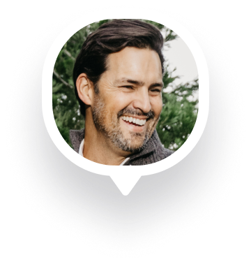 Smiling man with dark hair and beard outdoors with green foliage background.