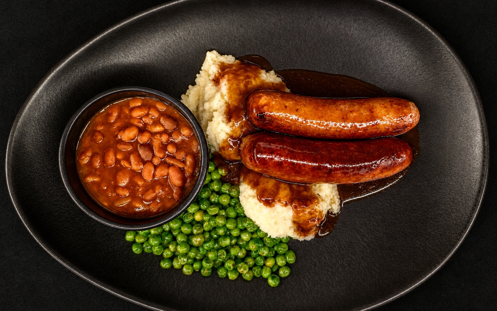 Bangers and mash with green peas, gravy and a side of baked beans