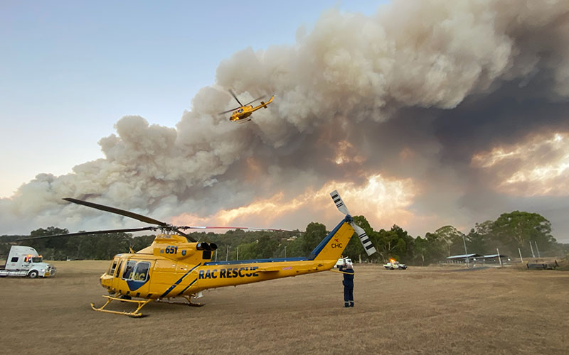 RAC Rescue Helicopter is landed in the foreground, one flies above, with a bushfire burning in the distance
