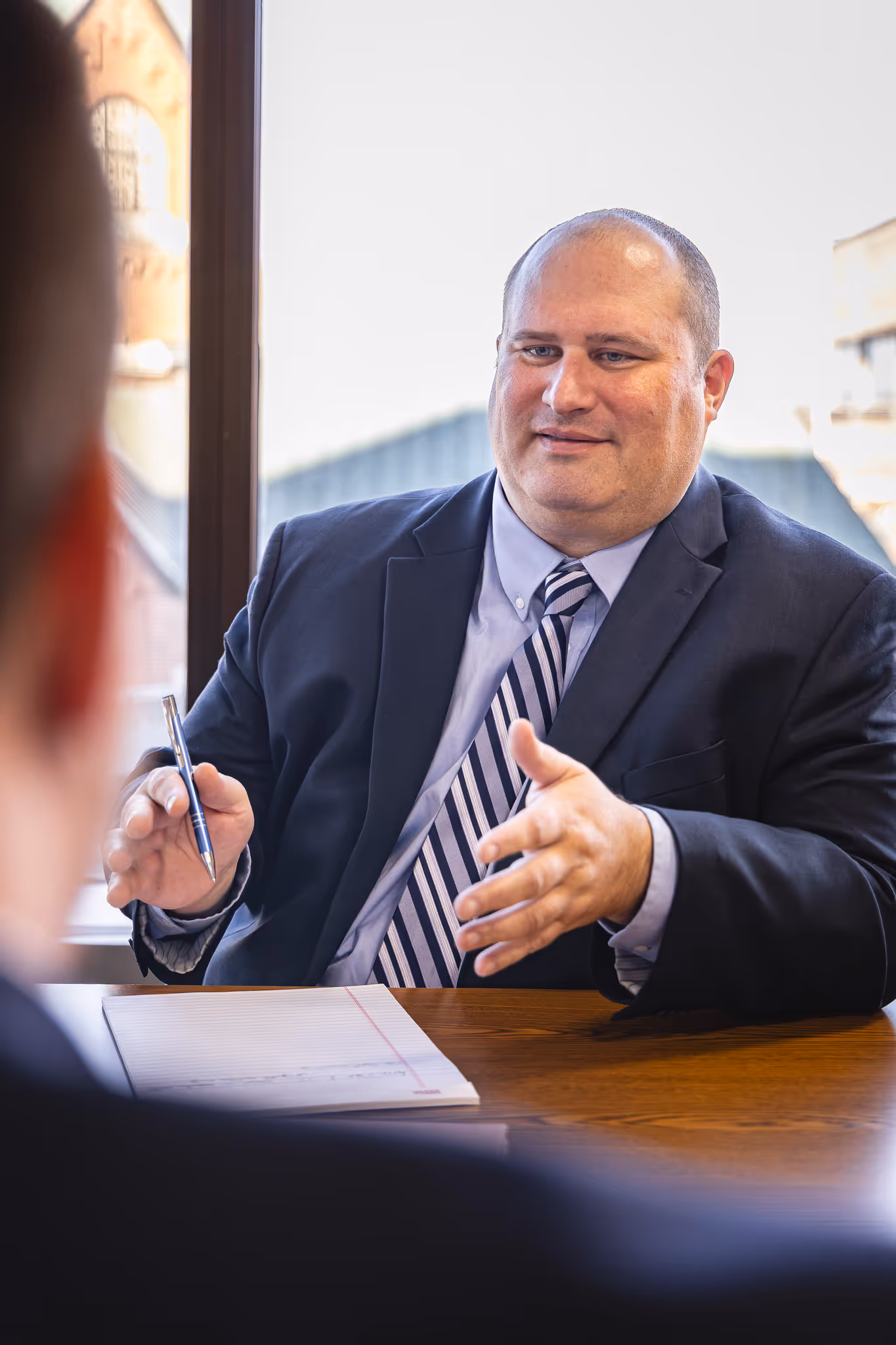 Man in suit and striped tie gesturing with one hand while holding a pen, seated at a desk with a notepad, speaking to another person.