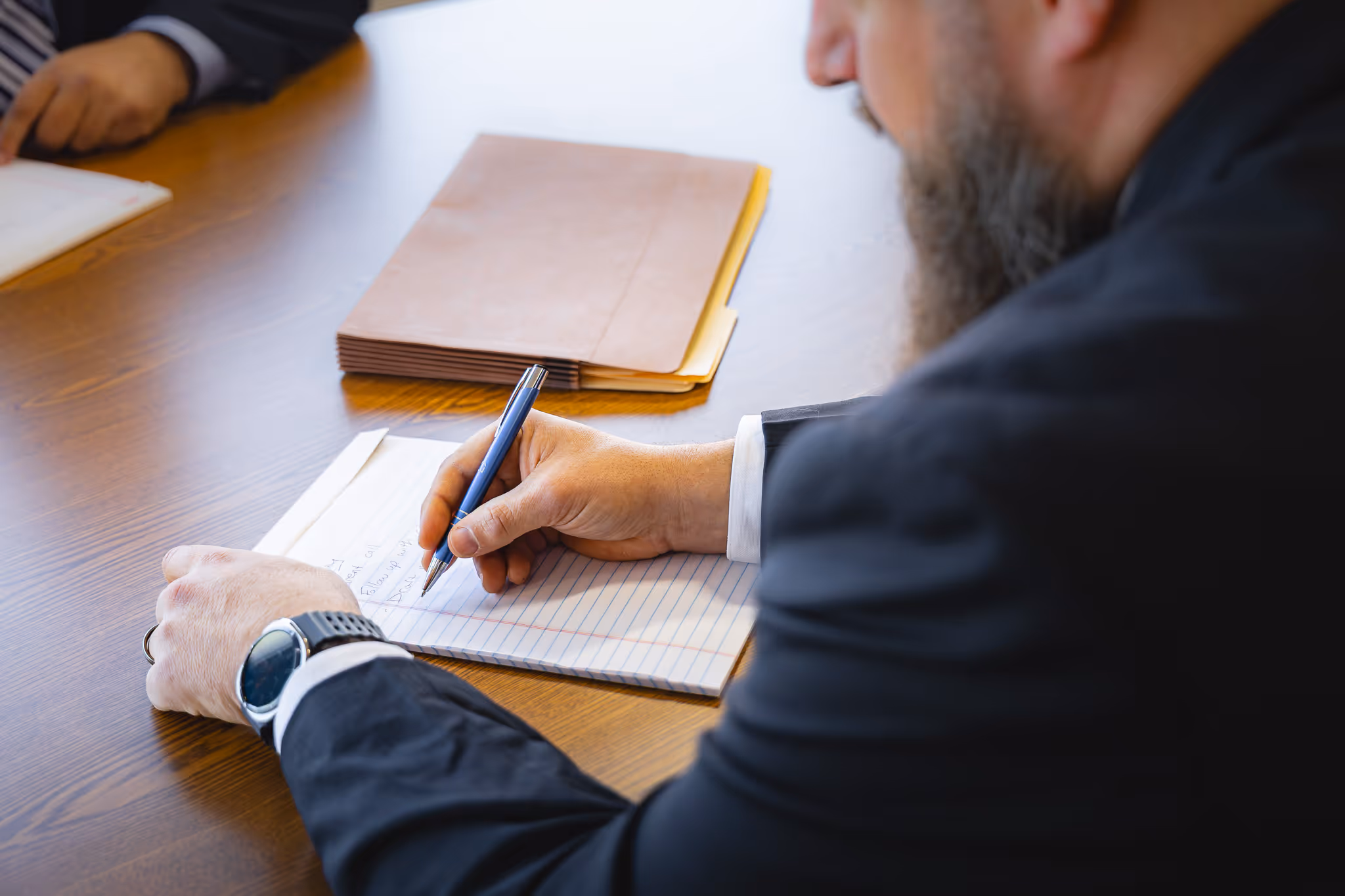 Man in business attire writing notes with a pen on lined paper at a wooden table with a closed folder.