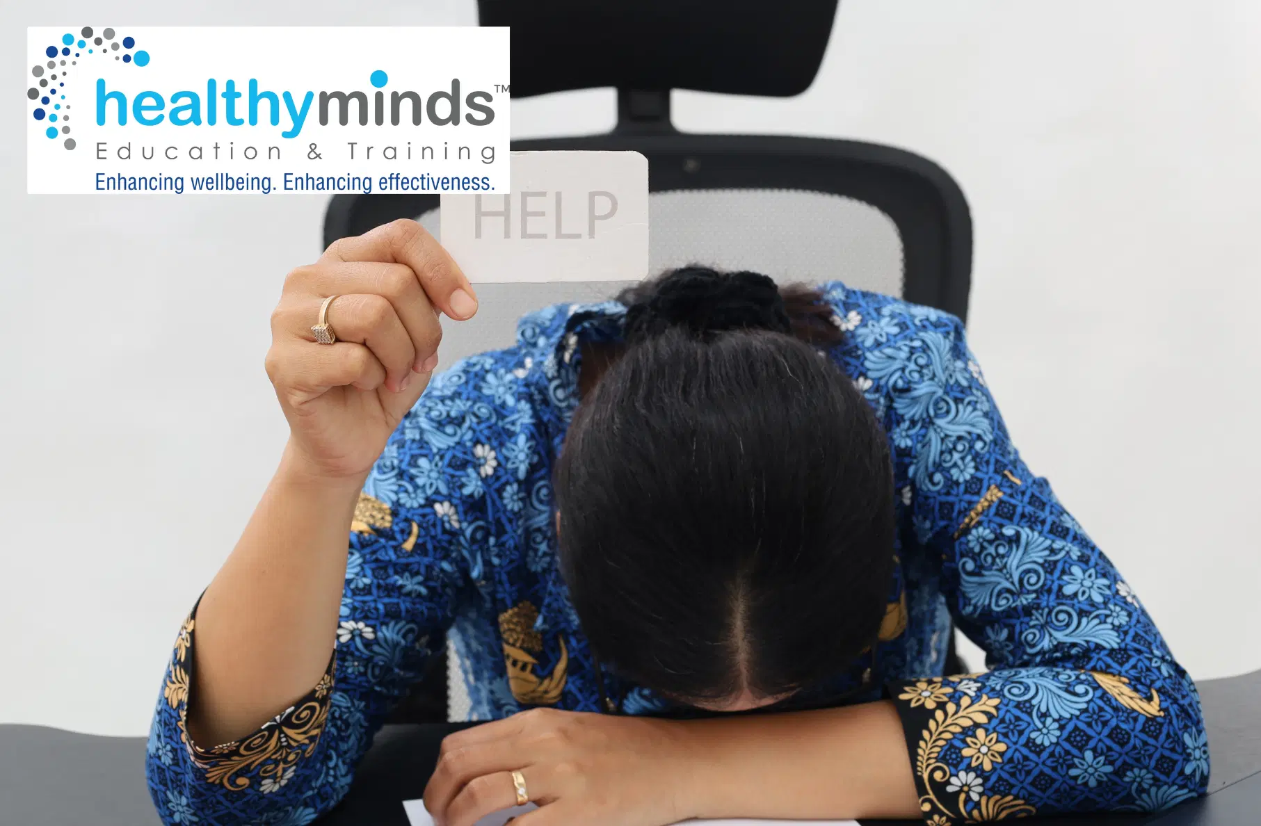 Person with head down on a desk holding a card that says HELP, symbolizing stress or need for support.