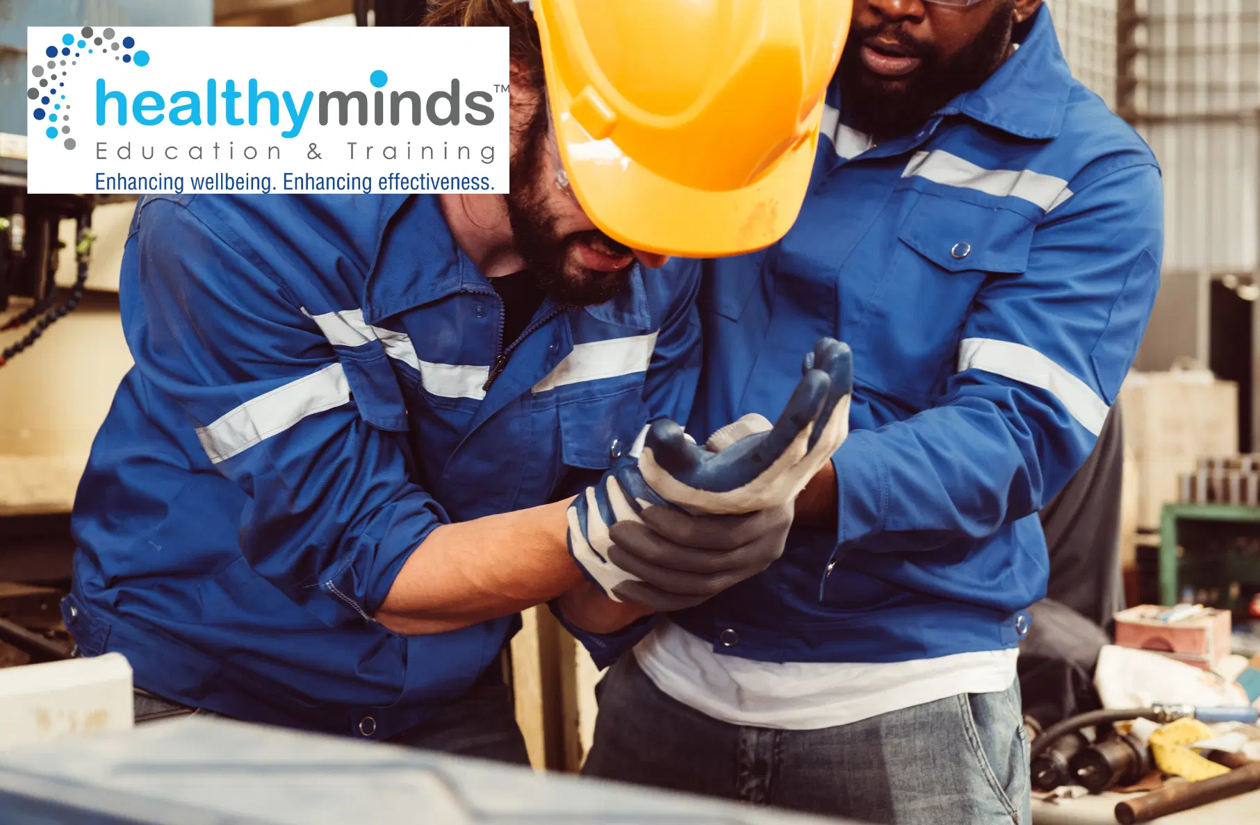 Construction worker in a yellow hard hat clutching his wrist in pain while being supported by a colleague in a workshop.