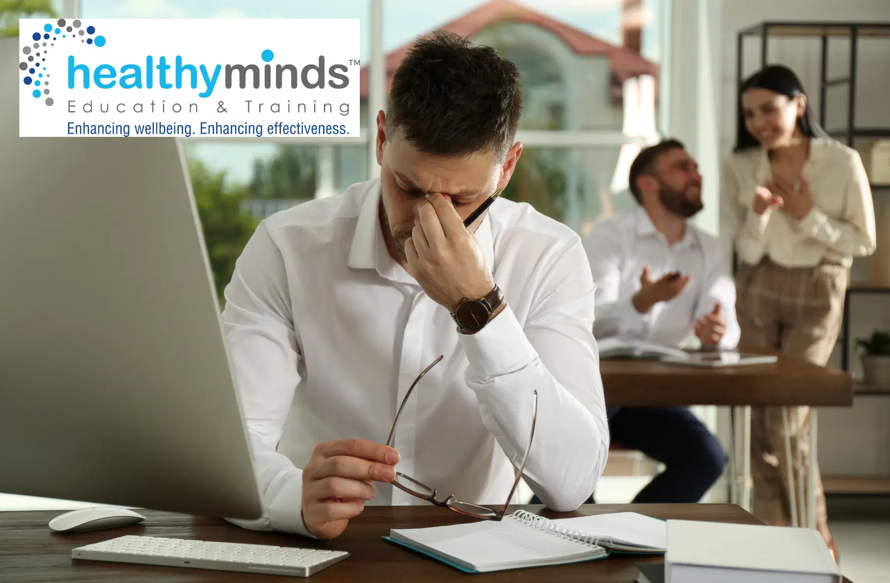 Stressed man in white shirt rubbing his eyes and holding glasses at desk while colleagues talk in background.