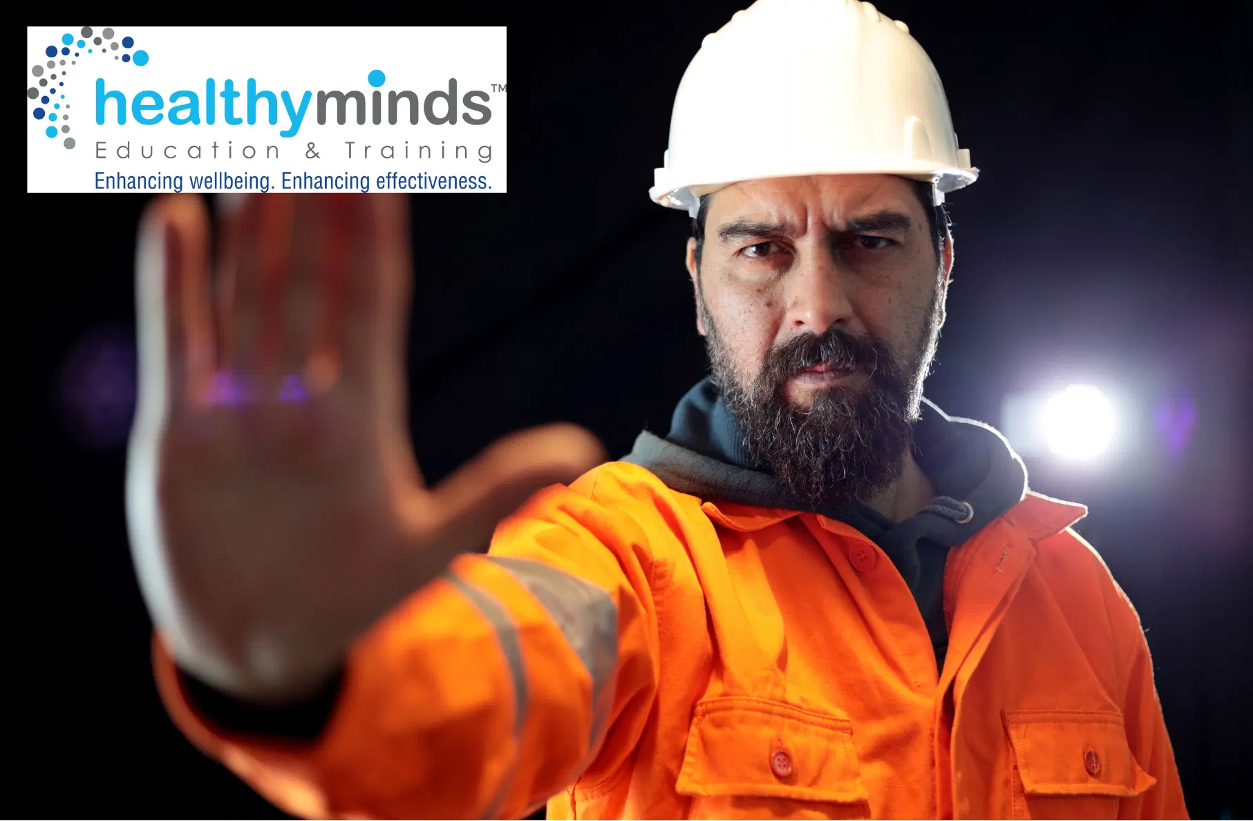 Serious construction worker in orange safety jacket and white hard hat raising his hand as a stop gesture against a dark background.