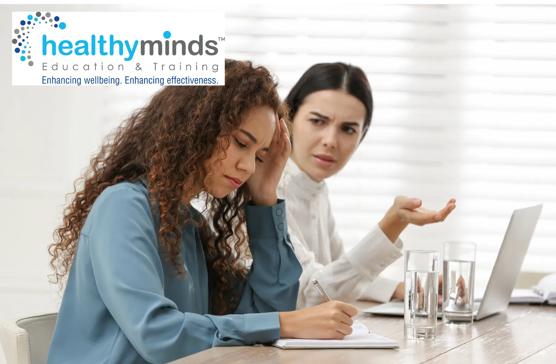Two women in a discussion at a desk, one looks stressed holding her head while the other gestures and appears concerned.