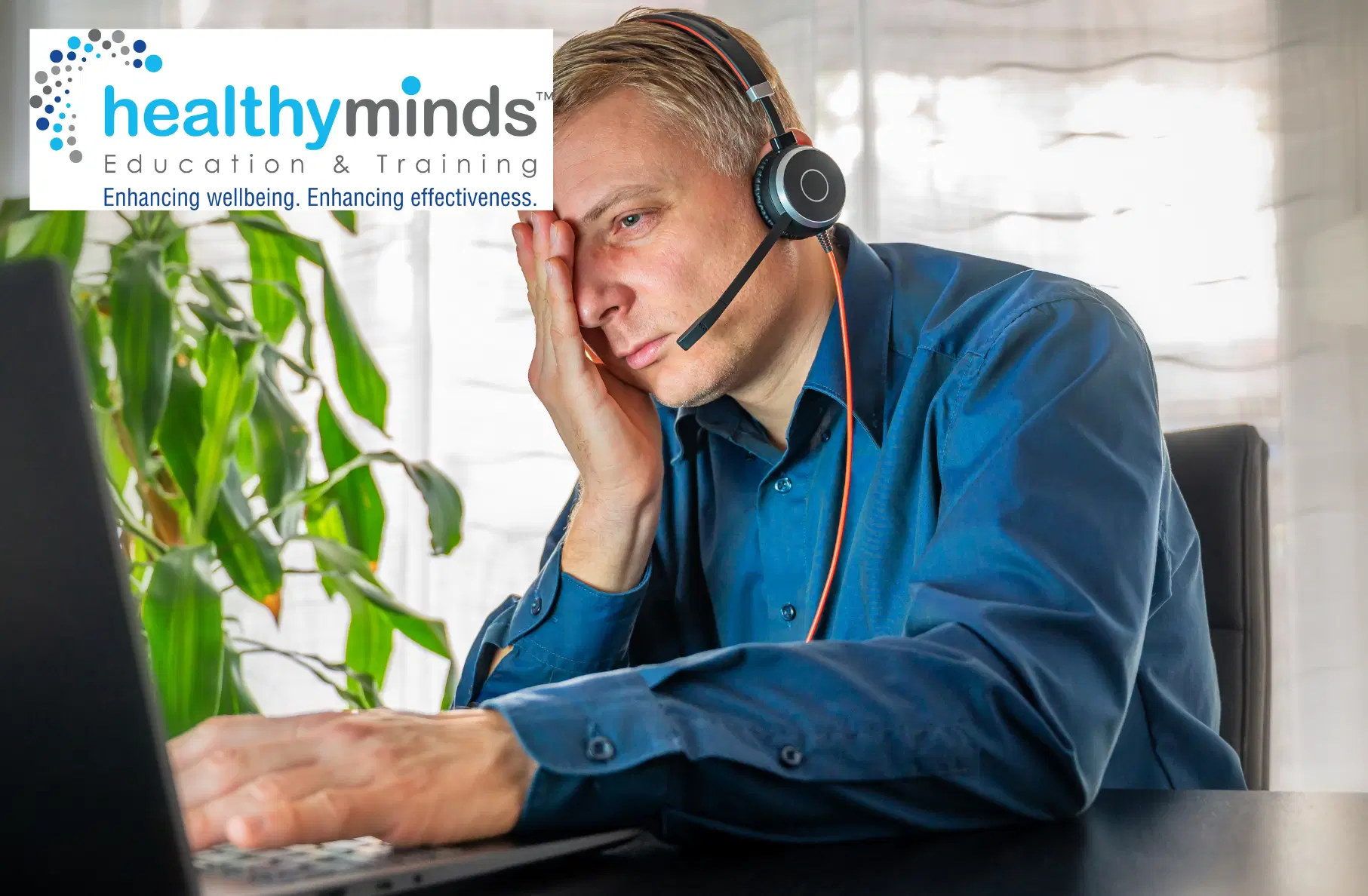 Stressed man wearing headset works on a laptop, resting his face on his hand.