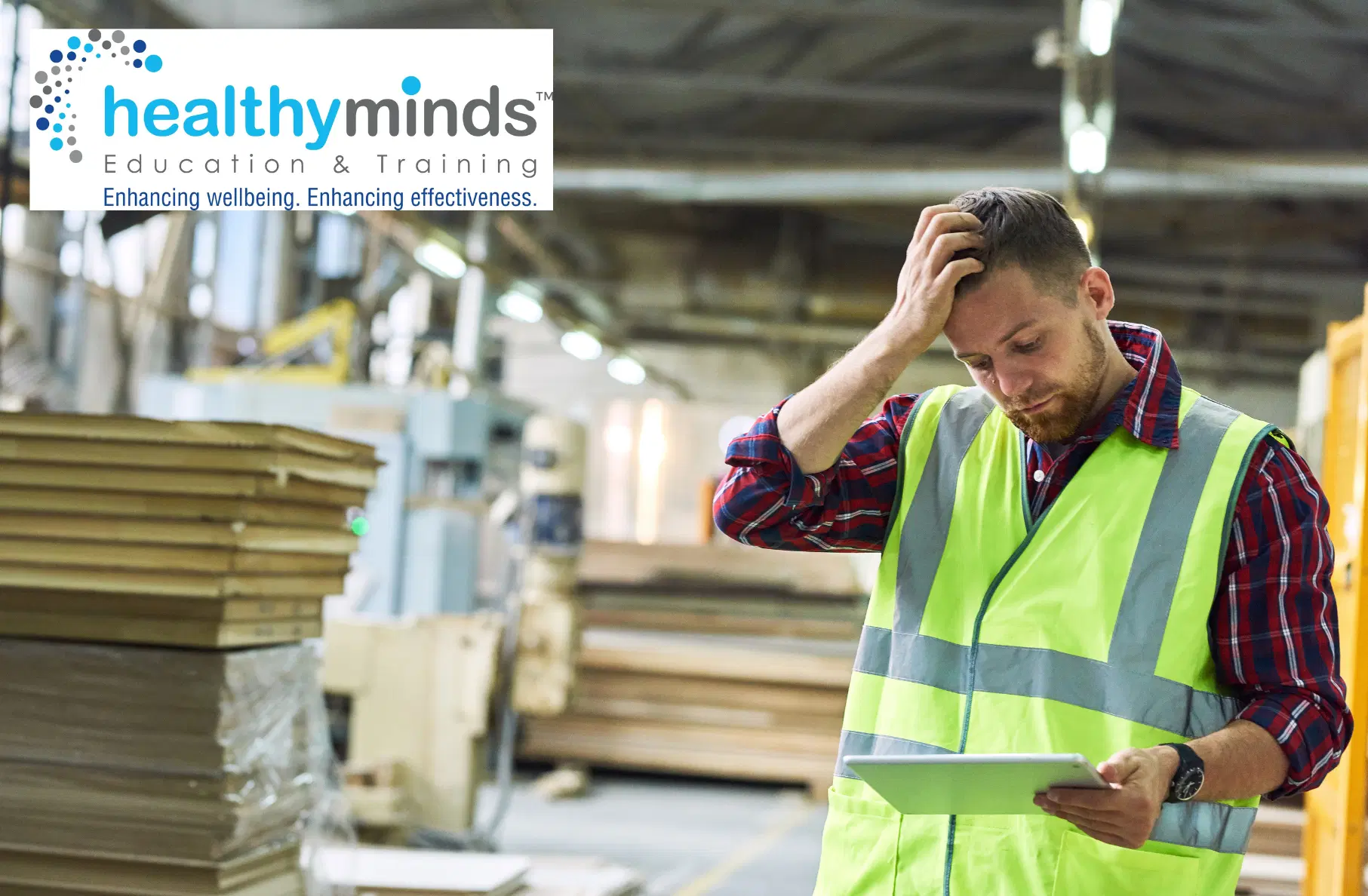 Worker in a neon safety vest and plaid shirt in a warehouse, looking confused while holding a tablet and scratching his head.