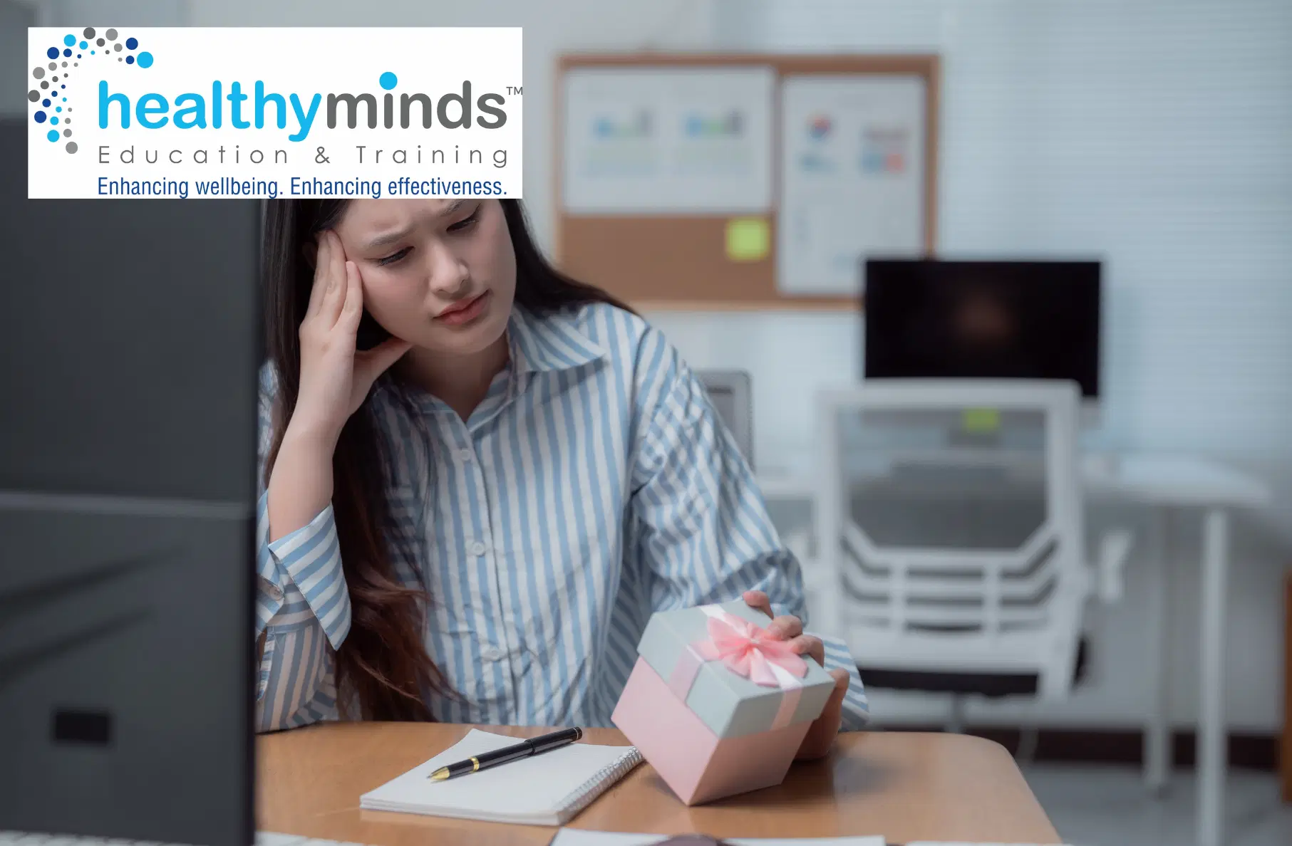 A woman in a striped shirt sitting at a desk, looking stressed while holding a small gift box with a pink ribbon.