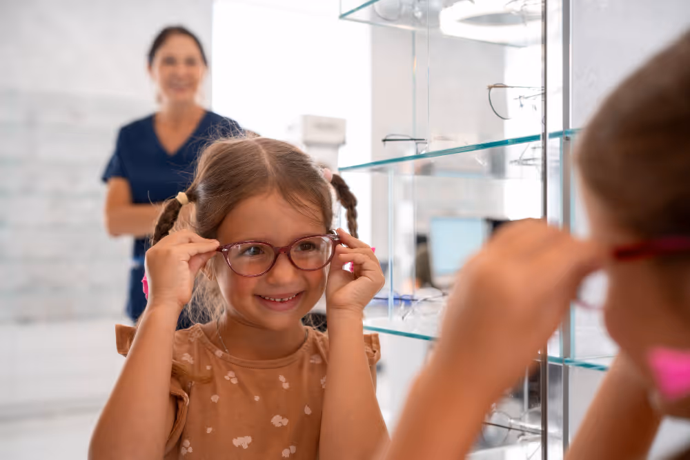 Smiling girl trying on glasses in optician's office with nurse behind