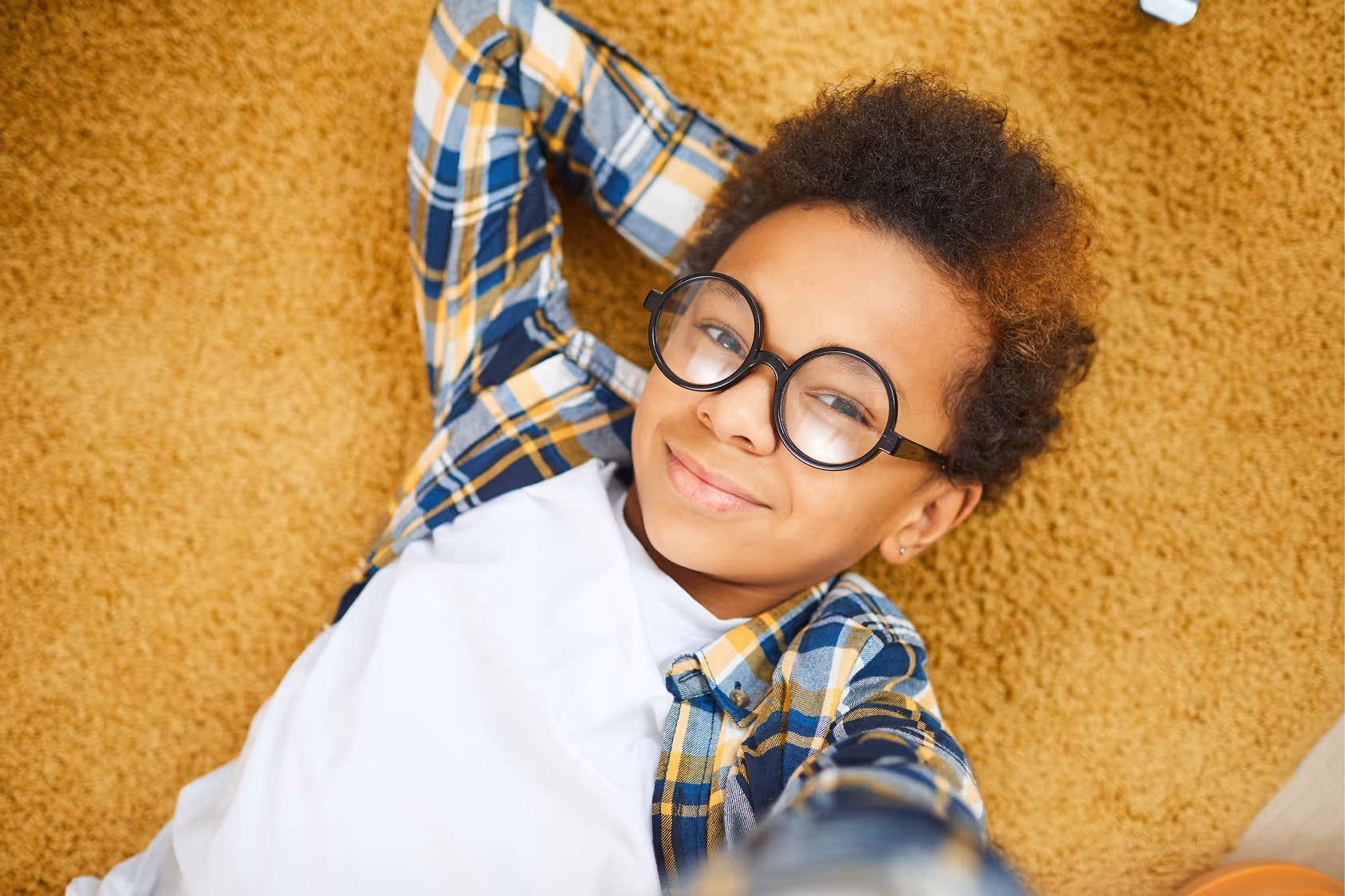Child in round glasses and plaid shirt lying on soft carpet smiling