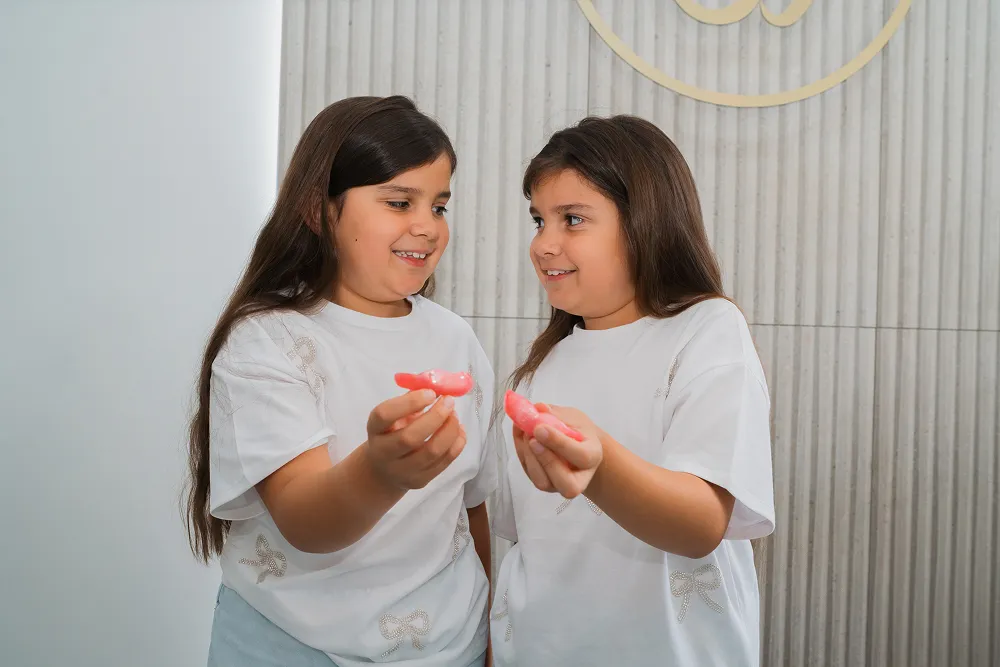 Two young girls in white shirts holding pink dental molds and smiling at each other.
