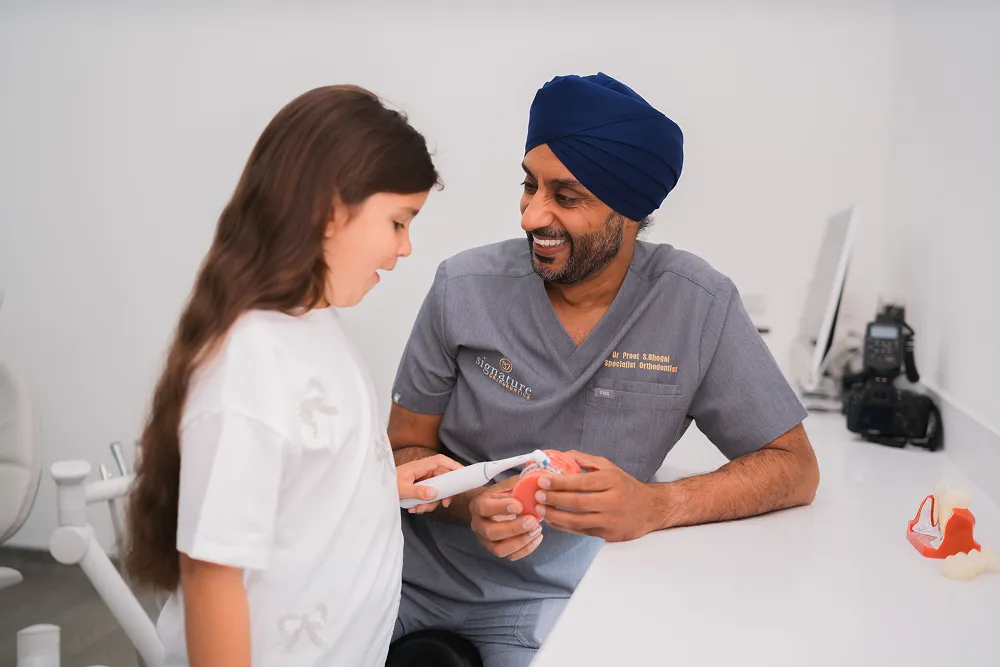 Orthodontist wearing a navy turban demonstrating teeth cleaning on a dental model to a young girl.