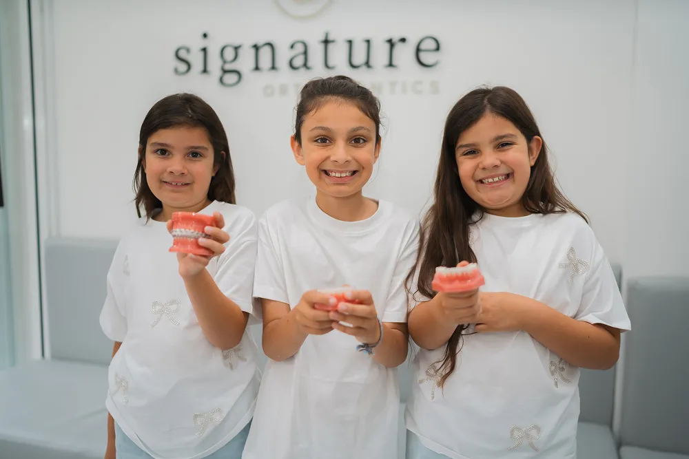 Three smiling girls in white shirts holding dental models at Signature Orthodontics.