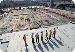 Construction site with workers in safety gear standing on a concrete platform overlooking rebar grid and foundation work.