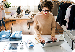 Person with curly hair and glasses working on a laptop at a retail counter with folded jeans and clothes racks nearby.