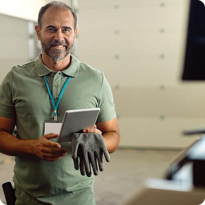 Smiling man wearing a green polo shirt and ID badge holds a tablet and a gray work glove in a bright workshop.