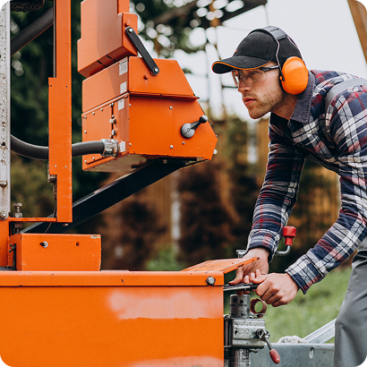 Man wearing ear protection and a cap operating an orange industrial machine outdoors.