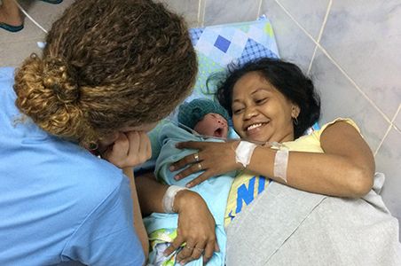 A mother joyfully holding her newborn while a supporter offers encouragement and care beside the hospital bed.