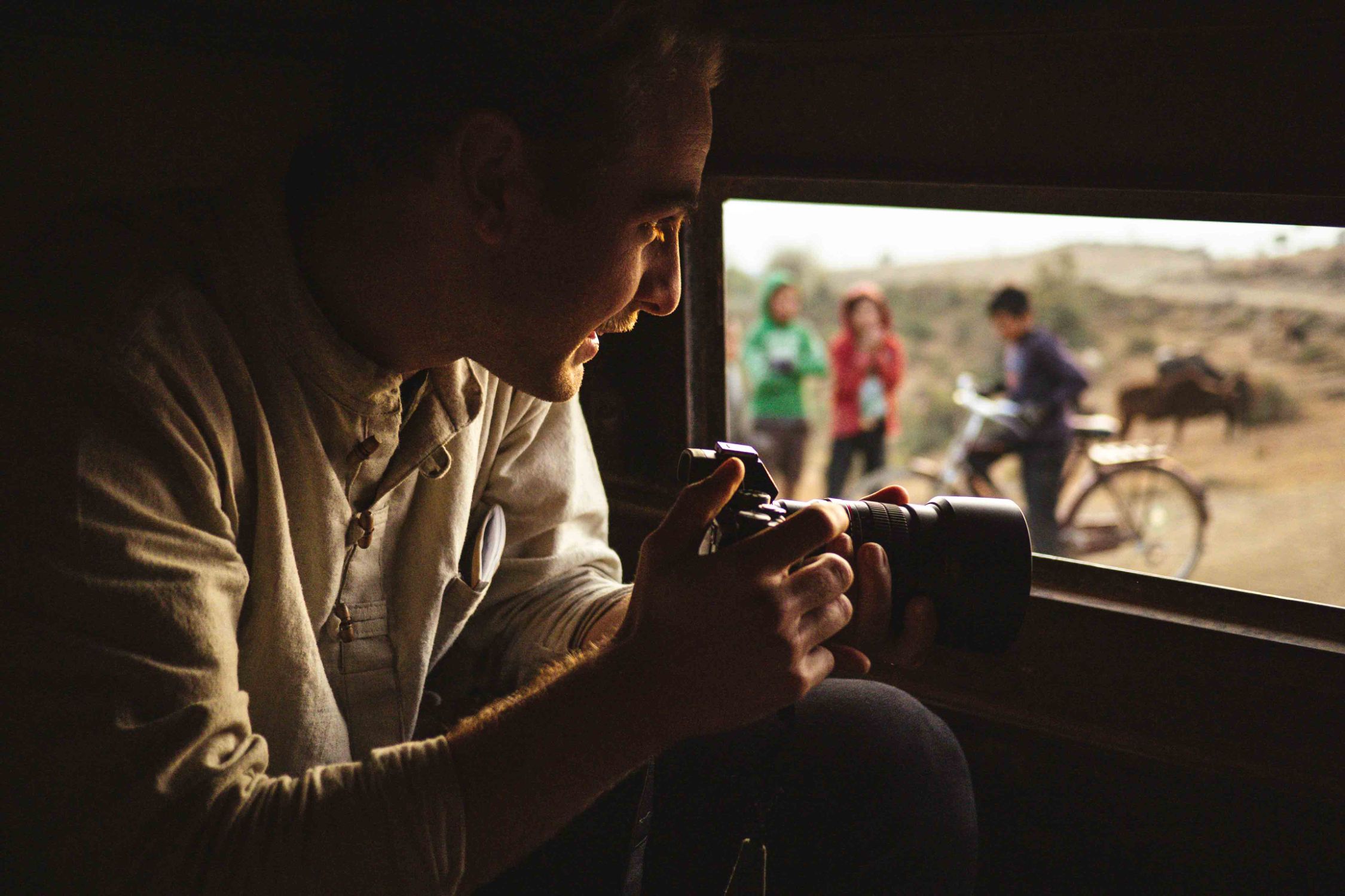 A filmmaker focuses his camera through a window, framing a distant moment of daily life unfolding outside.