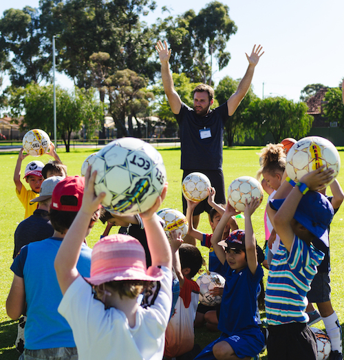 A coach leads a group of children holding soccer balls during a sunny outdoor sports session.